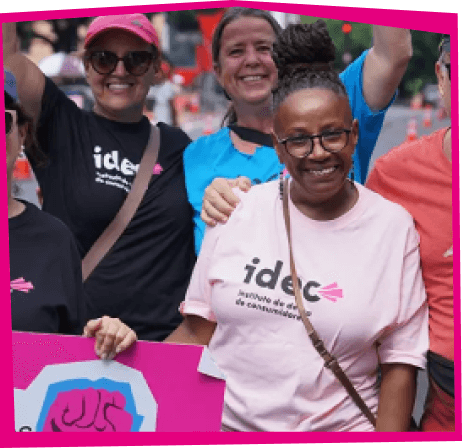 Foto de um grupo de seis pessoas sorrindo e abraçadas, todas vestindo camisetas do IDEC. As camisetas têm várias cores, incluindo preto, rosa e amarelo. Eles estão participando de um evento ao ar livre