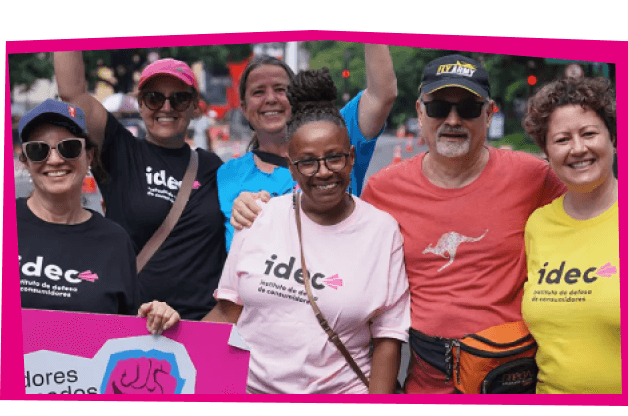 Foto de um grupo de seis pessoas sorrindo e abraçadas, todas vestindo camisetas do IDEC. As camisetas têm várias cores, incluindo preto, rosa e amarelo. Eles estão participando de um evento ao ar livre